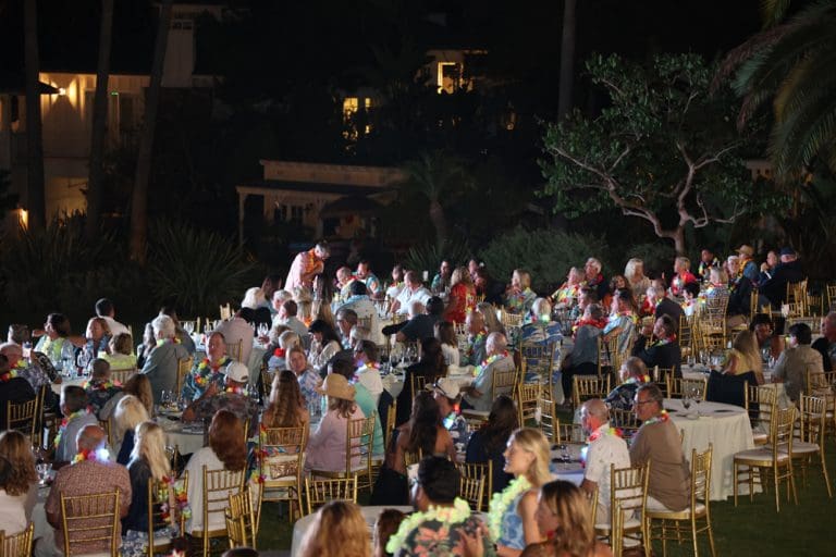 A large group of people seated at outdoor round tables at night, wearing leis and surrounded by greenery, enjoy a lively Catalinaville-themed event.