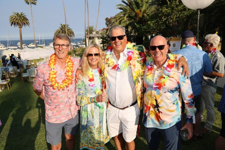 Four adults in colorful, tropical-themed outfits and leis pose together outdoors at Catalinaville, a sunny event with palm trees and boats in the background.