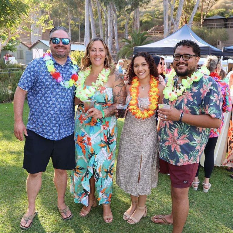 Four adults wearing leis and summer outfits stand together outdoors on grass, smiling at the camera during Catalinaville, a daytime hospital fundraising event with trees, tents, and record-breaking energy in the background.
