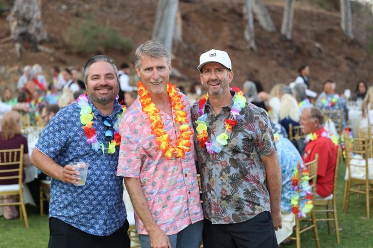 Three men wearing Hawaiian shirts and leis pose and smile at the Catalinaville outdoor event, with tables and people in the background.